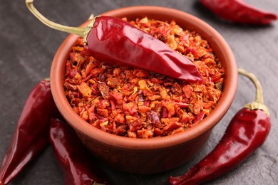 Chili pepper flakes in bowl and pods on dark textured table, closeup