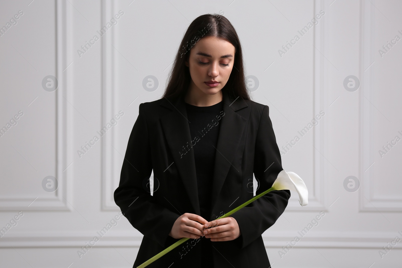 Photo of Sad woman with calla lily flower near white wall. Funeral ceremony