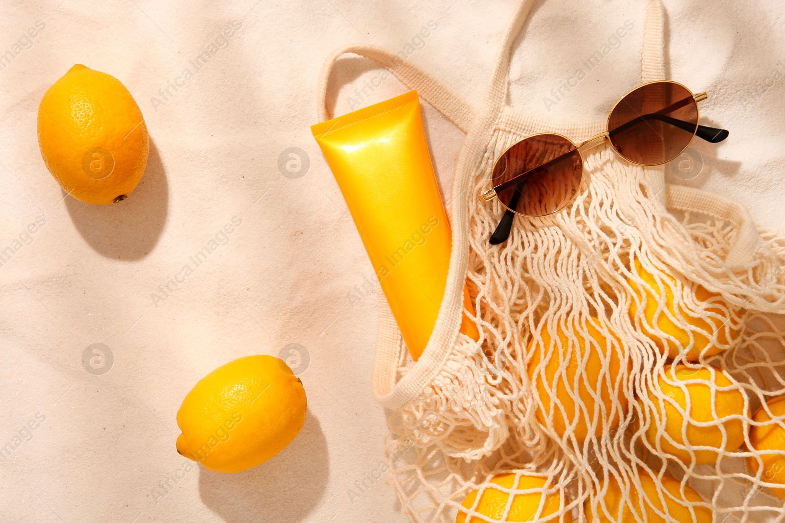 Photo of String bag with sunglasses, lemons and sunscreen on sand, flat lay