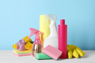 Photo of Spring cleaning. Detergents, flowers and sponges on white wooden table against light blue background