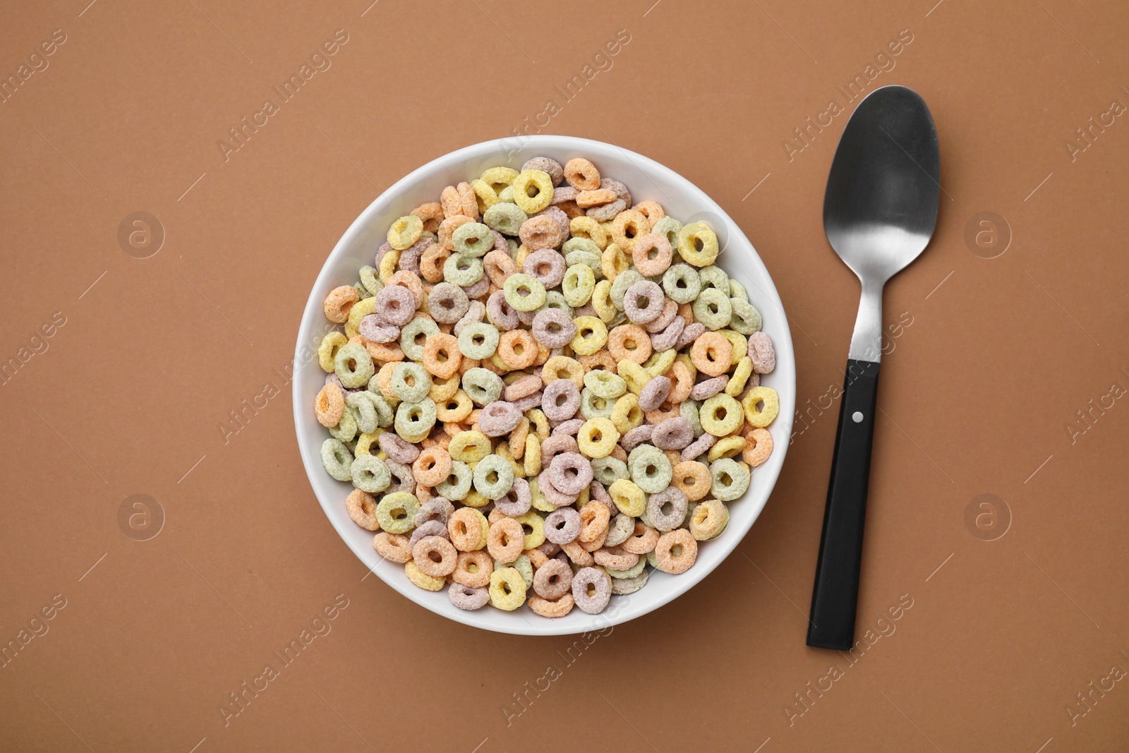 Photo of Tasty cereal rings in bowl and spoon on brown table, flat lay