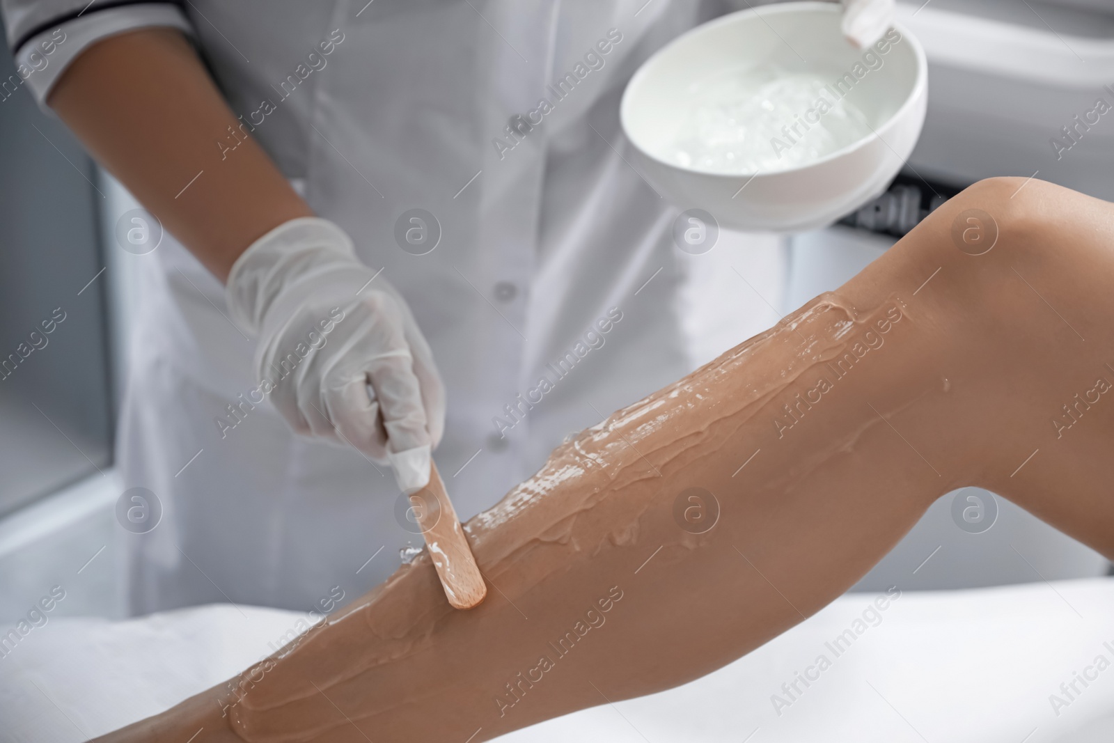 Photo of Professional cosmetologist applying gel on client's leg before laser epilation procedure in salon, closeup