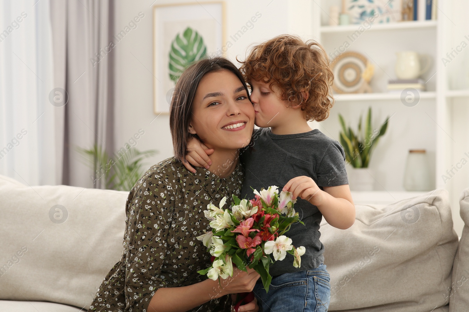 Photo of Little son kissing and congratulating his mom with Mother`s day at home. Woman holding bouquet of flowers