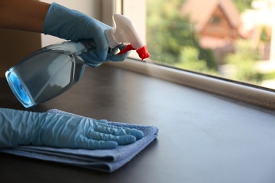 Photo of Woman in gloves cleaning grey stone window sill with rag and detergent indoors, closeup
