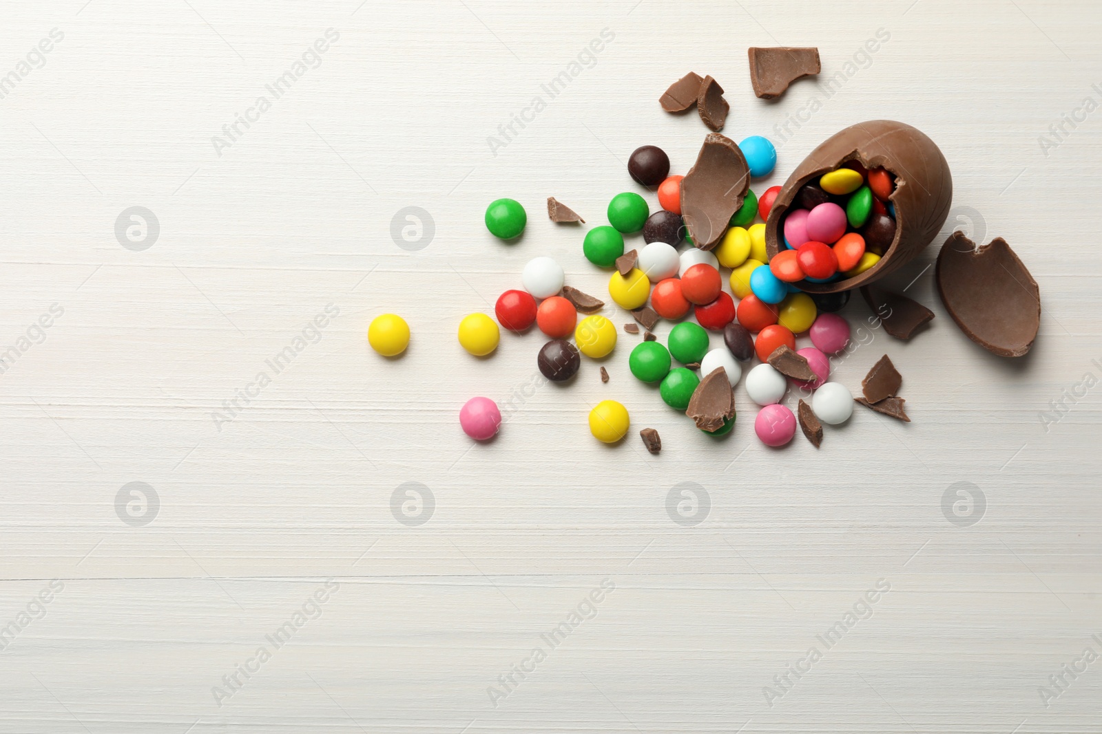 Photo of Broken chocolate egg and colorful candies on white wooden table, flat lay. Space for text