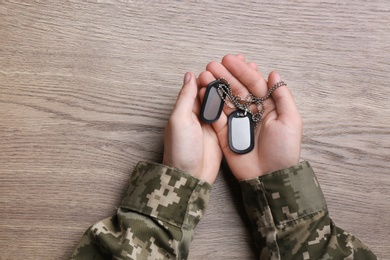 Photo of Man in camouflage uniform holding military ID tags over wooden background, top view