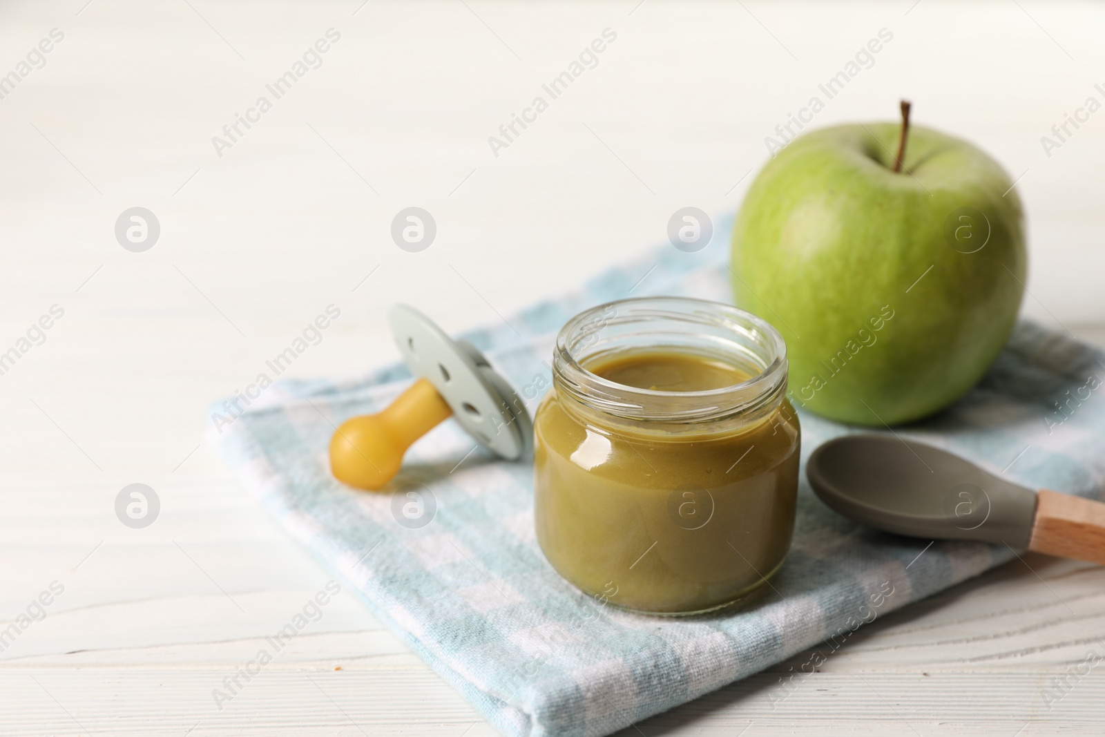 Photo of Jar with healthy baby food, apple, pacifier and spoon on white wooden table. Space for text