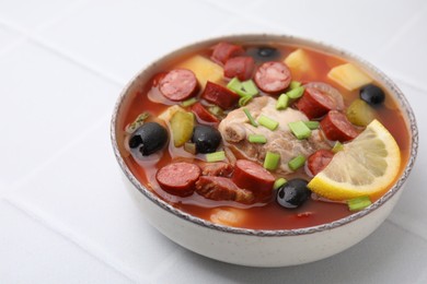 Meat solyanka soup with thin dry smoked sausages in bowl on white tiled table, closeup. Space for text