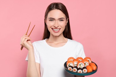 Photo of Happy young woman with plate of sushi rolls and chopsticks on pink background
