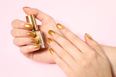 Woman holding bottle of golden nail polish in manicured hand on color background, closeup