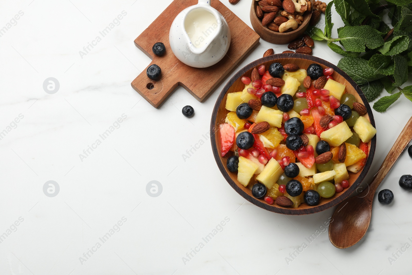 Photo of Delicious fruit salad in bowl, berries, fresh mint and nuts on white marble table, flat lay. Space for text
