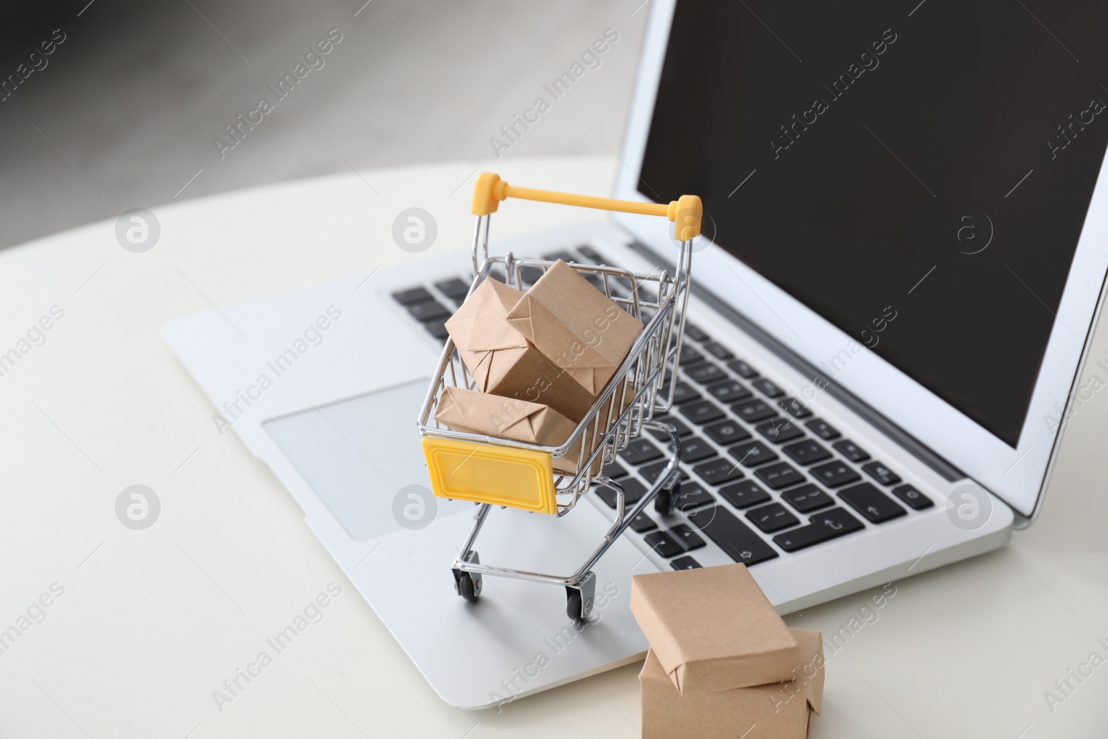 Photo of Internet shopping. Laptop and small cart with boxes on white table indoors