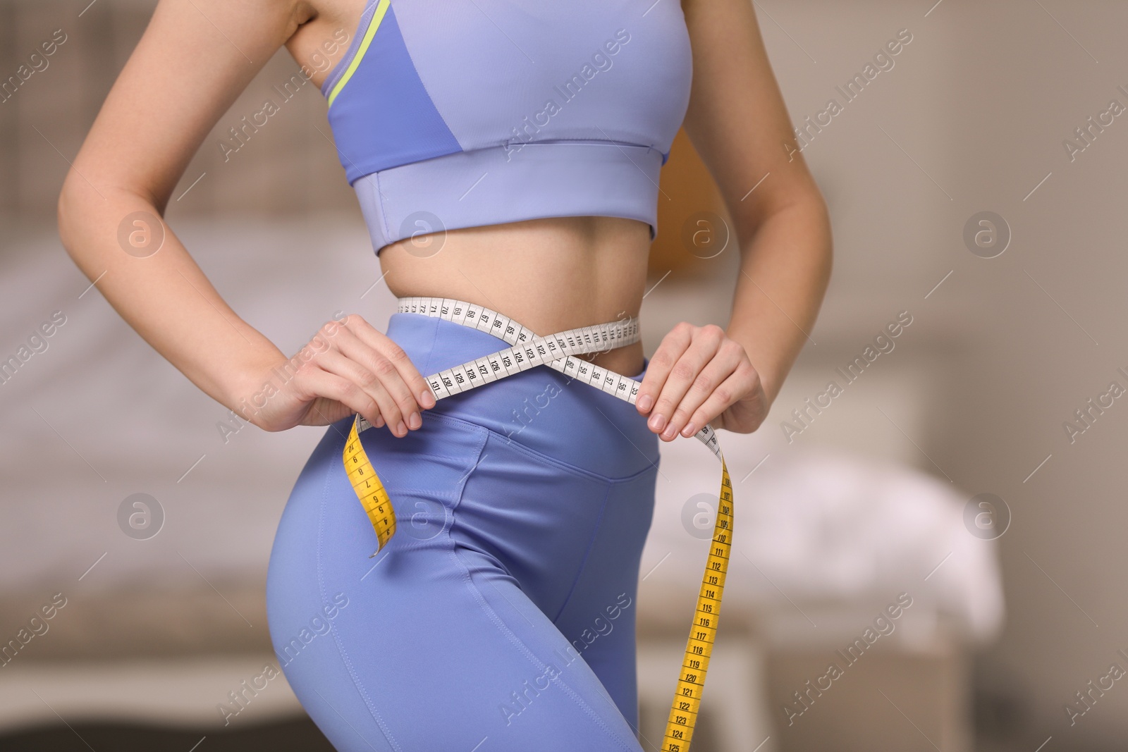 Photo of Woman in sportswear measuring waist with tape at home, closeup