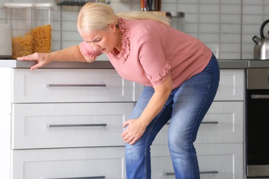 Senior woman suffering from knee pain in kitchen