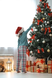 Photo of Cute little child in Santa hat decorating Christmas tree at home