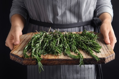 Woman holding wooden board with fresh rosemary twigs on black background, closeup