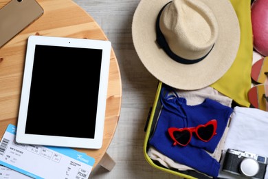 Modern tablet, tickets and smartphone on wooden table near packed suitcase, top view. Summer vacation