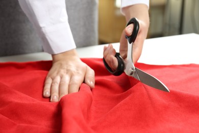 Photo of Seamstress cutting red fabric with scissors at workplace, closeup