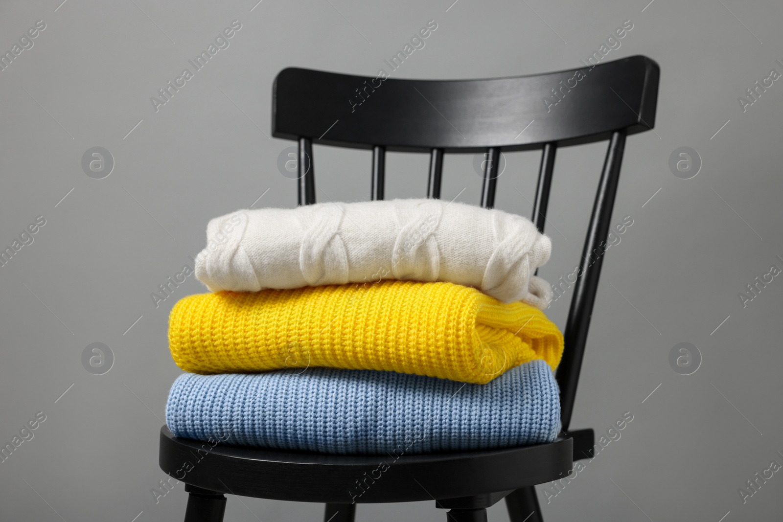 Photo of Stack of different casual sweaters on black chair against grey background