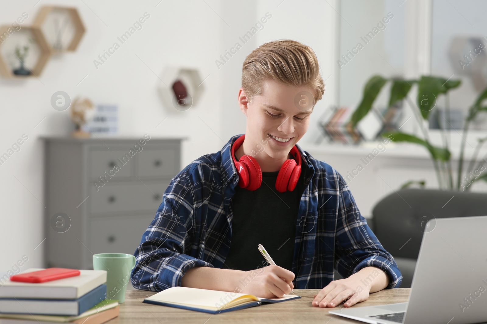 Photo of Online learning. Smiling teenage boy writing in notebook near laptop at home