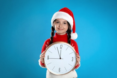 Girl in Santa hat with clock on light blue background. New Year countdown