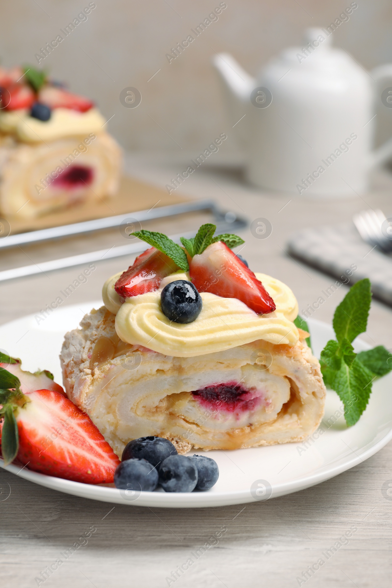 Photo of Piece of tasty meringue roll with jam, cream, strawberry, blueberry and mint on white wooden table, closeup