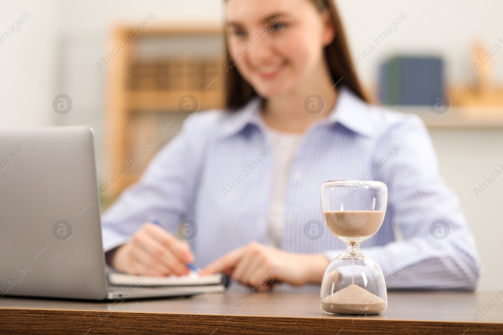 Photo of Hourglass with flowing sand on desk. Woman taking notes while using laptop indoors, selective focus