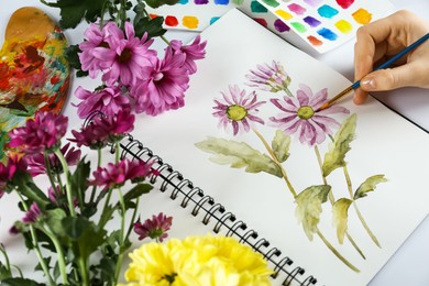 Photo of Woman painting chrysanthemums in sketchbook and flowers at white table, closeup