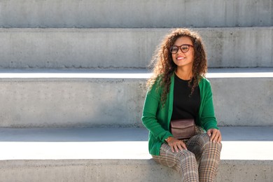 Beautiful African American woman with stylish waist bag on stairs outdoors, space for text