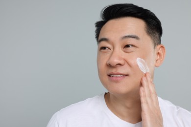 Handsome man applying cream onto his face on light grey background. Space for text