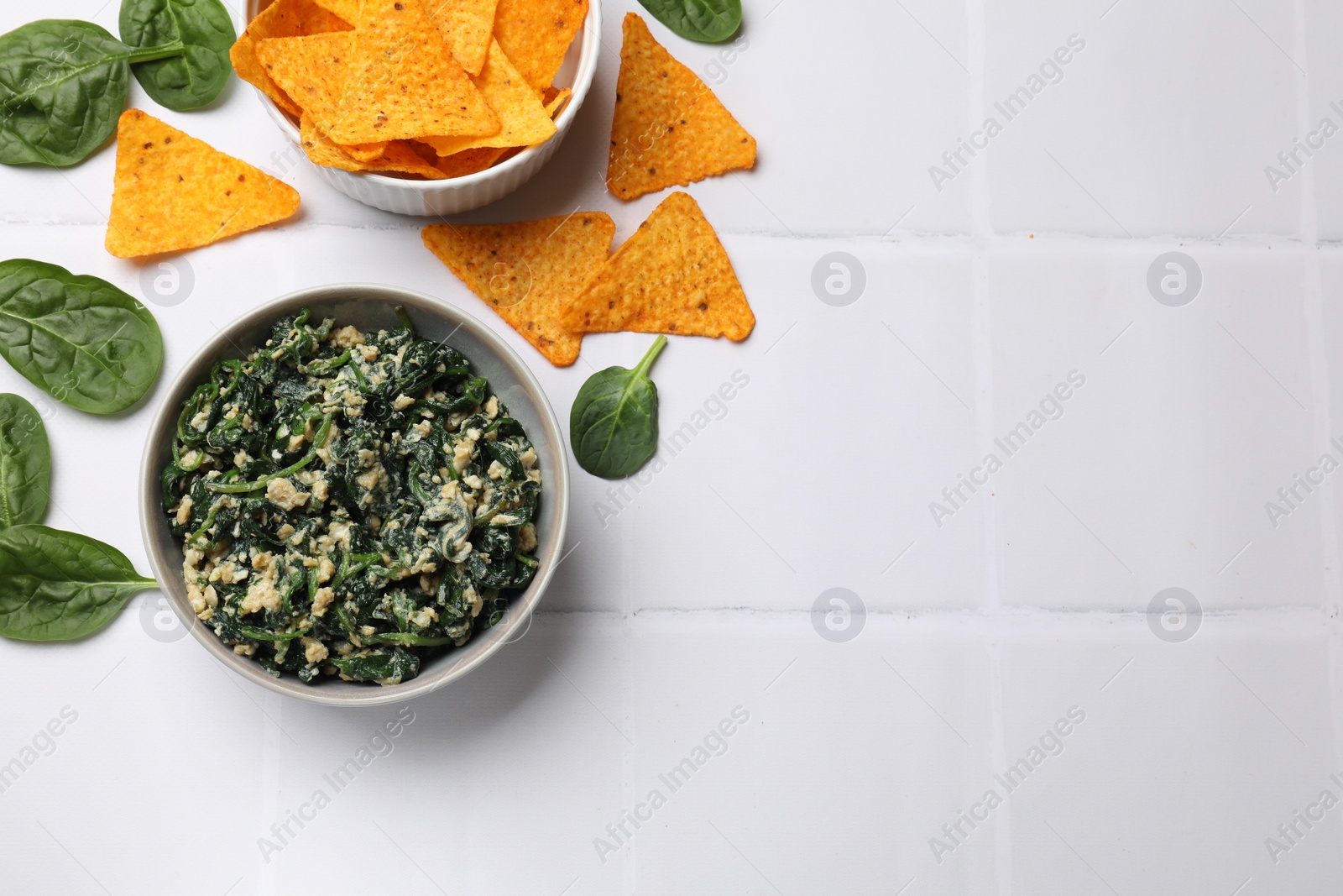 Photo of Tasty spinach dip with egg in bowl and nachos chips on white tiled table, flat lay. Space for text