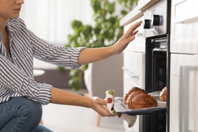 Beautiful young woman taking out tray of baked buns from oven in kitchen