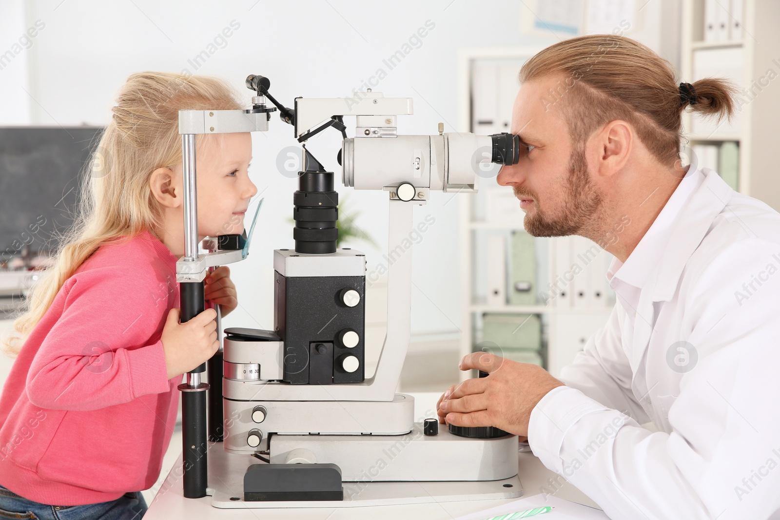 Photo of Children's doctor examining little girl with ophthalmic equipment in clinic