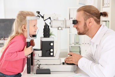 Photo of Children's doctor examining little girl with ophthalmic equipment in clinic