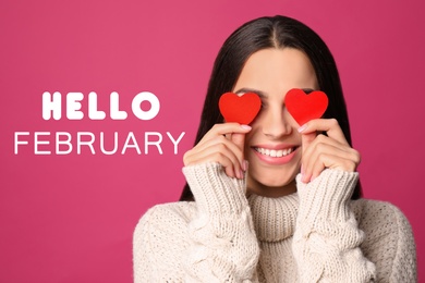 Greeting card with text Hello February. Young woman holding paper hearts near eyes on pink background