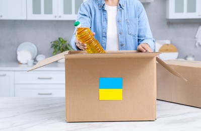 Humanitarian aid for Ukraine. Woman putting bottle of oil in donation box at white table indoors, closeup