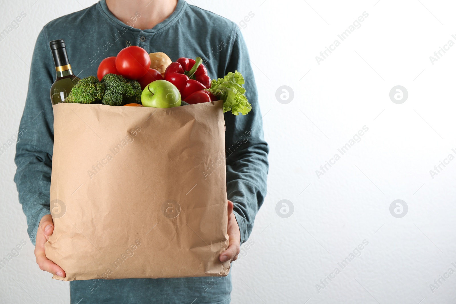 Photo of Man holding paper bag with different groceries near white wall, closeup view. Space for text