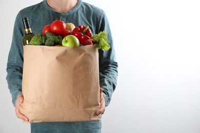 Man holding paper bag with different groceries near white wall, closeup view. Space for text