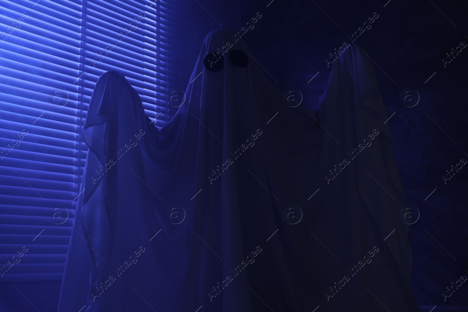 Photo of Creepy ghost. Woman covered with sheet near window in blue light