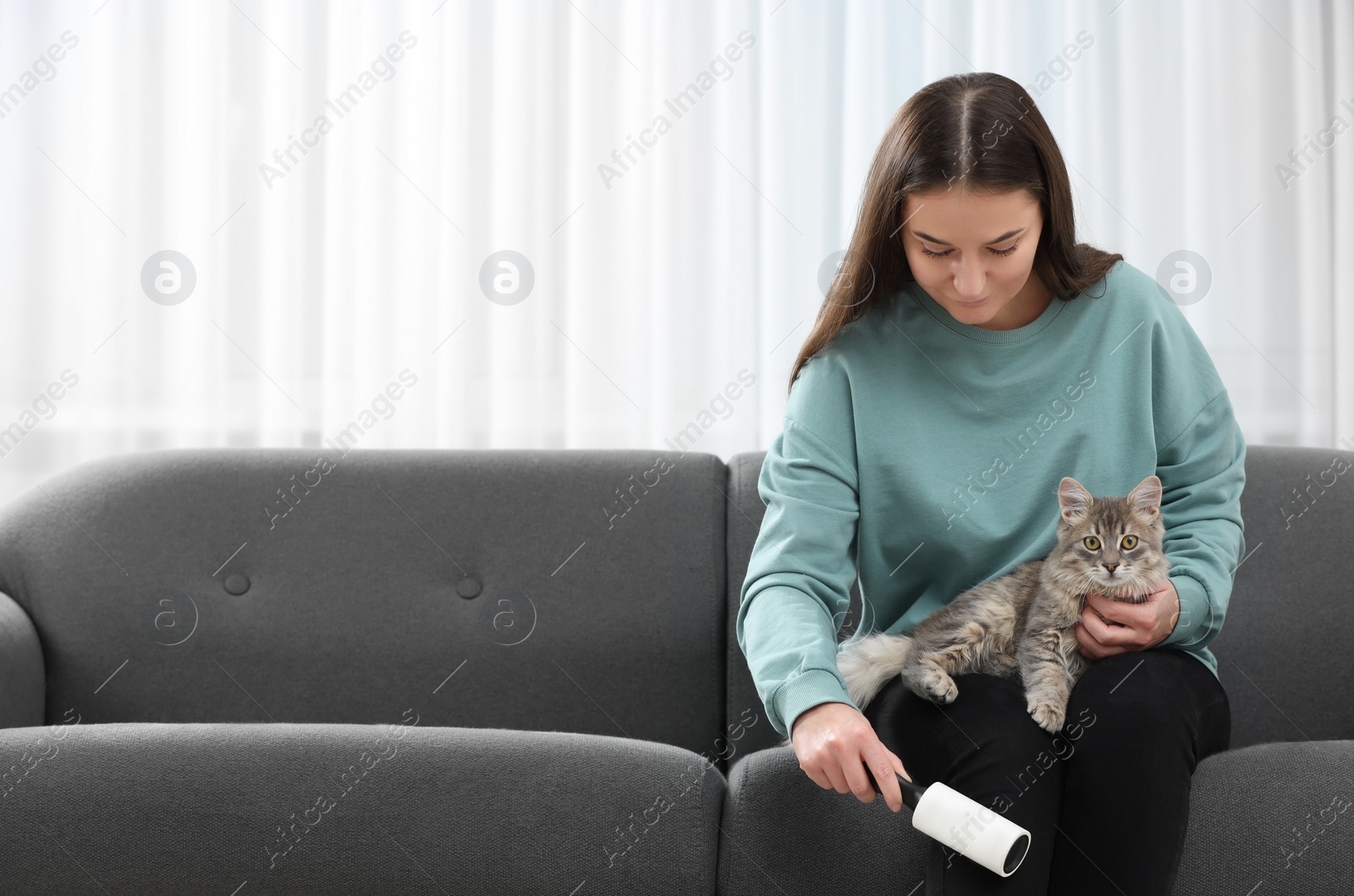 Photo of Pet shedding. Woman with lint roller removing cat`s hair from trousers on sofa at home, space for text