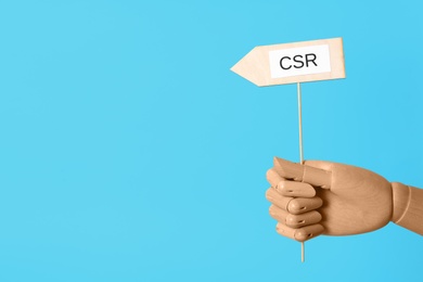 Photo of Wooden mannequin holding sign with abbreviation CSR on light blue background, closeup. Corporate social responsibility