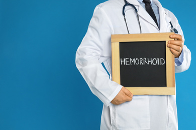 Photo of Doctor holding blackboard with word HEMORRHOID on blue background, closeup. Space for text