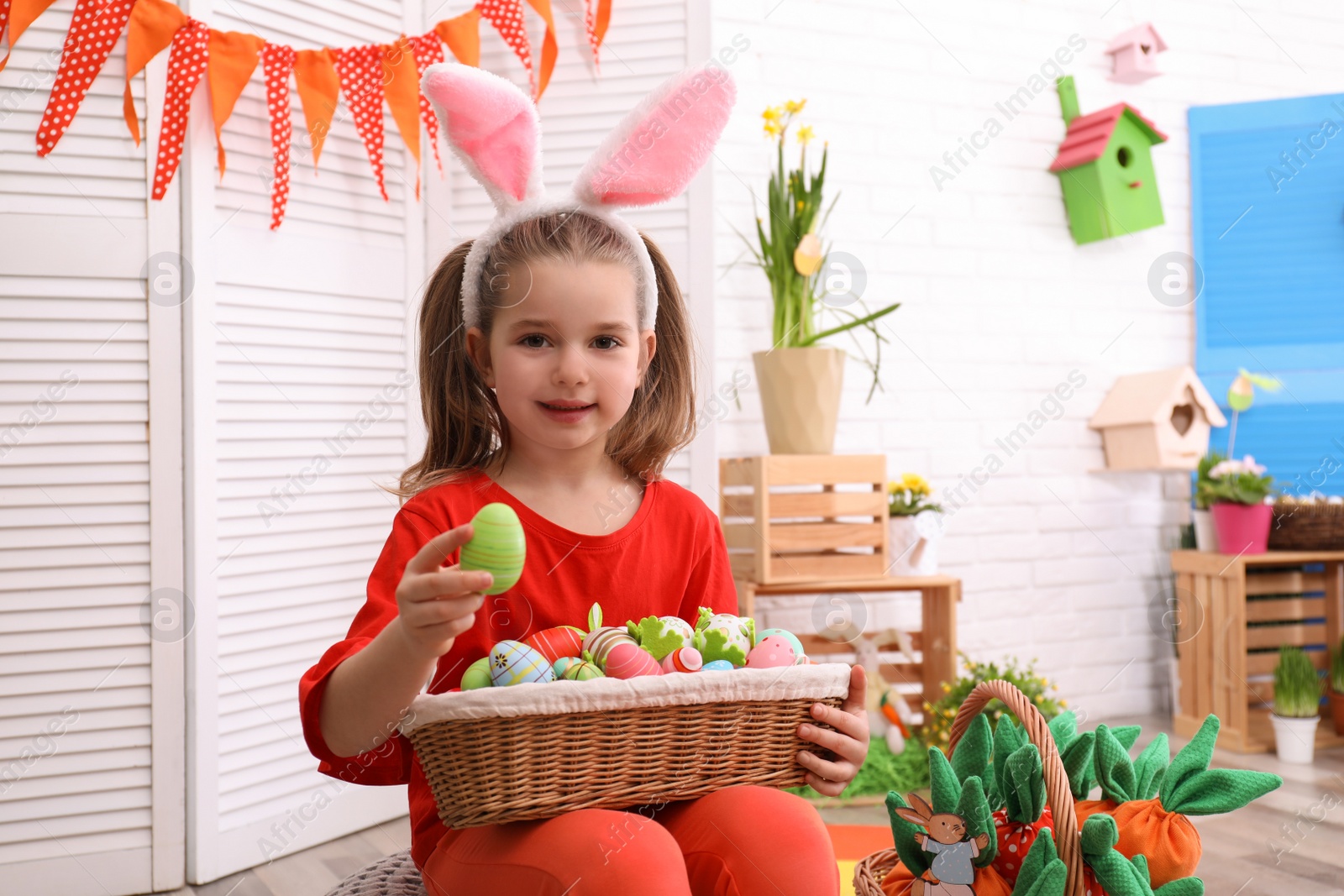 Photo of Happy little girl with bunny ears and wicker basket full of Easter eggs indoors
