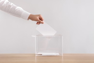 Photo of Woman putting her vote into ballot box on wooden table against light grey background, closeup