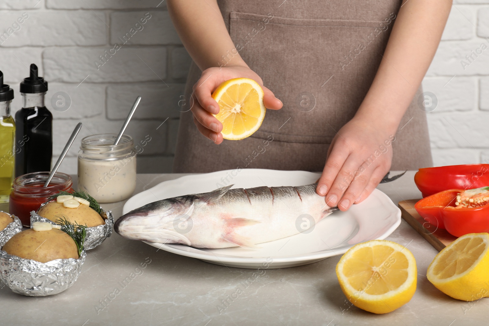 Photo of Woman squeezing lemon juice on fresh sea bass fish at light gray marble table, closeup