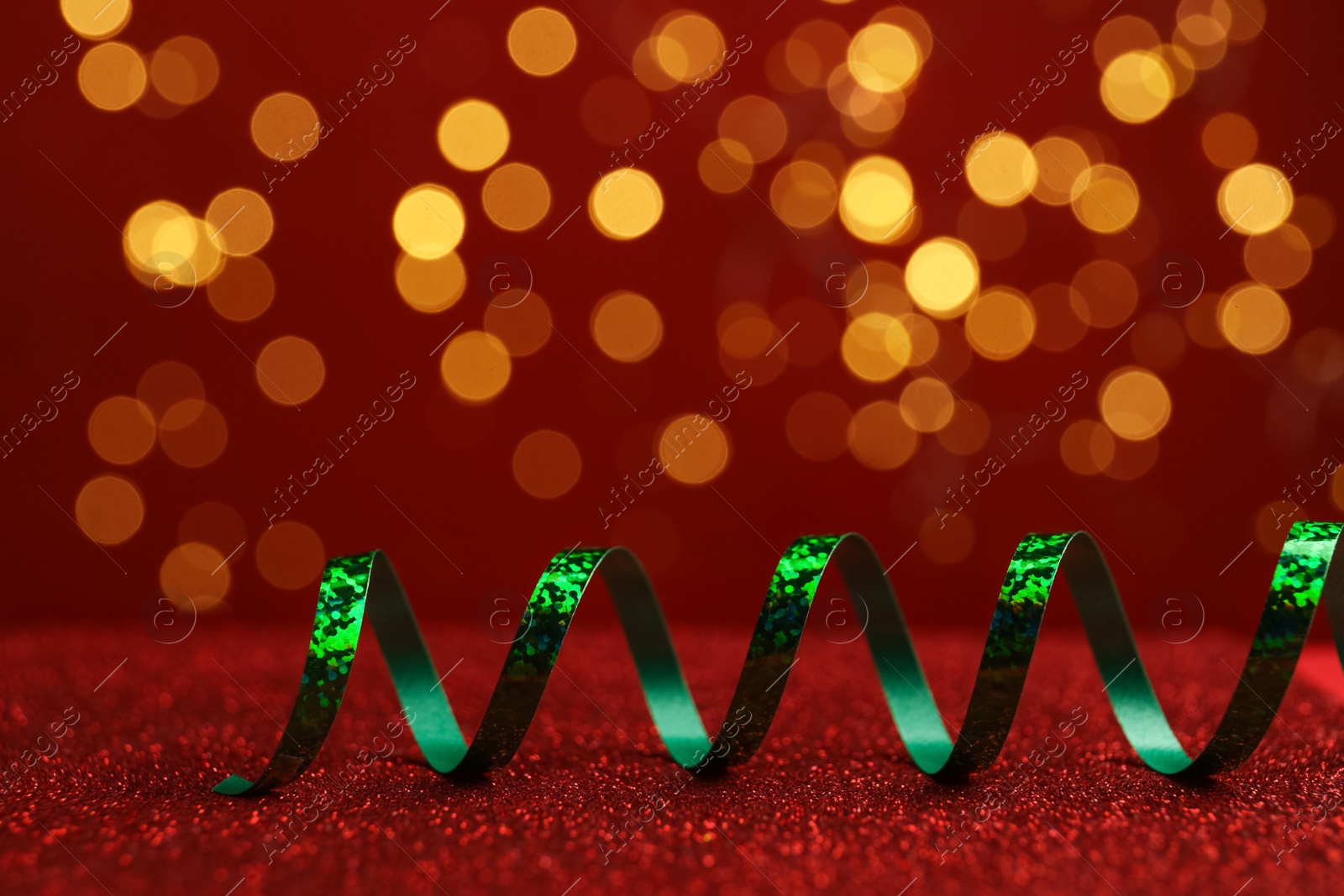 Photo of Shiny green serpentine streamer on red table against blurred lights. Space for text