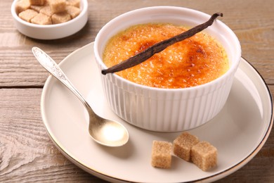 Delicious creme brulee in bowl, vanilla pod, spoon and sugar cubes on wooden table, closeup