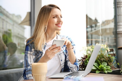 Photo of Young woman using mobile phone while working with laptop at desk in cafe