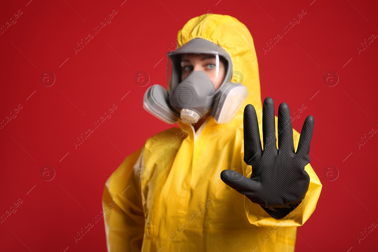 Photo of Woman in chemical protective suit making stop gesture against red background, focus on hand. Virus research