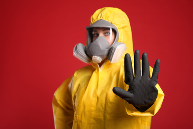 Photo of Woman in chemical protective suit making stop gesture against red background, focus on hand. Virus research
